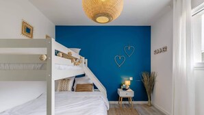 A bedroom with bunk beds, a blue accent wall, heart-shaped decorations, a bedside table with a lamp, and a window with curtains. - Clair Jeanne - Climatisation - Plage (Notre-Dame-de-Monts)