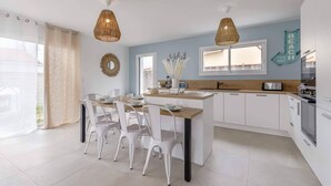 A modern kitchen with white cabinetry, a central island with chairs, pendant lights, and a view of the outside. - Clair Jeanne - Climatisation - Plage (Notre-Dame-de-Monts)