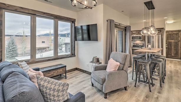 A spacious living room with a large window, a grey sofa, a grey armchair, and a dining area with stools and a table. - Sleek Columbia Falls Condo: Centrally Located! (Columbia Falls)
