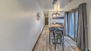 A hallway with a wooden floor, a dining area with a table and bar stools, a large window with curtains, and a painting on the wall. - Sleek Columbia Falls Condo: Centrally Located! (Columbia Falls)