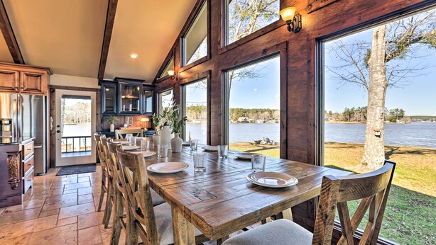 A wooden dining table set with plates and glasses, large windows with a view of a lake, and a kitchen area with wooden cabinets. - Lay Lake Access & Views: Expansive Shelby Home! (Shelby)
