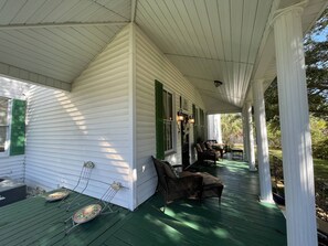 Relax on the covered porch with wicker chairs and a small table, surrounded by a lush green yard.