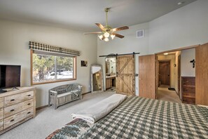 Quarto com cama, um banquinho, c ômoda, televisão, ventilador de teto e janela com vista para árvores e neve.