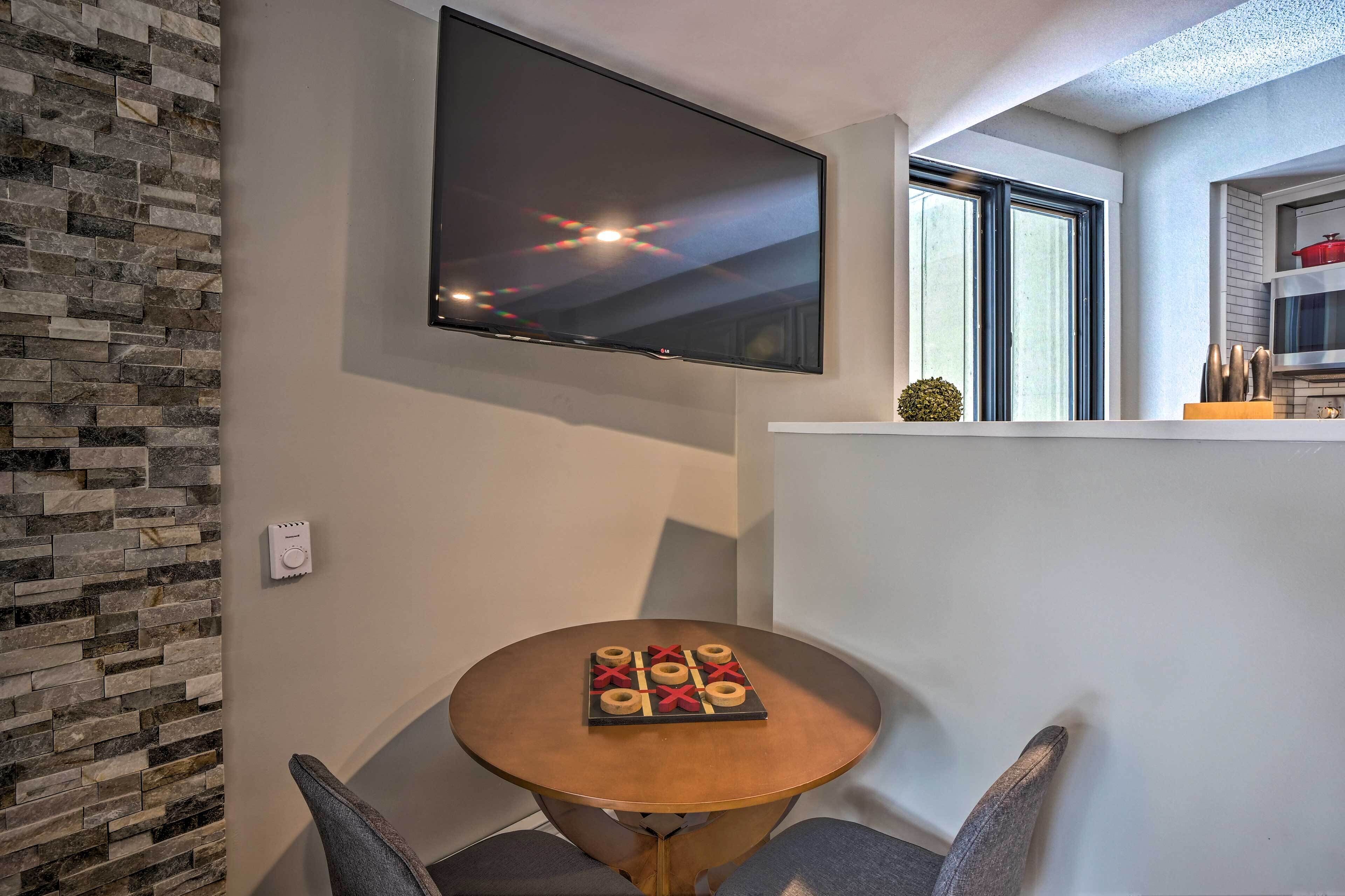 A modern living room with a flat-screen TV mounted on the wall, a round wooden table with a board game, and a stone-patterned wall.