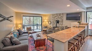 A modern living room with a large sofa, a coffee table, and a dining area with a long counter and bar stools. There is a TV mounted on the wall and a fireplace. - < 1 Mi to Peak 8: Ski Condo in Breck w/ Mtn Views (Breckenridge)
