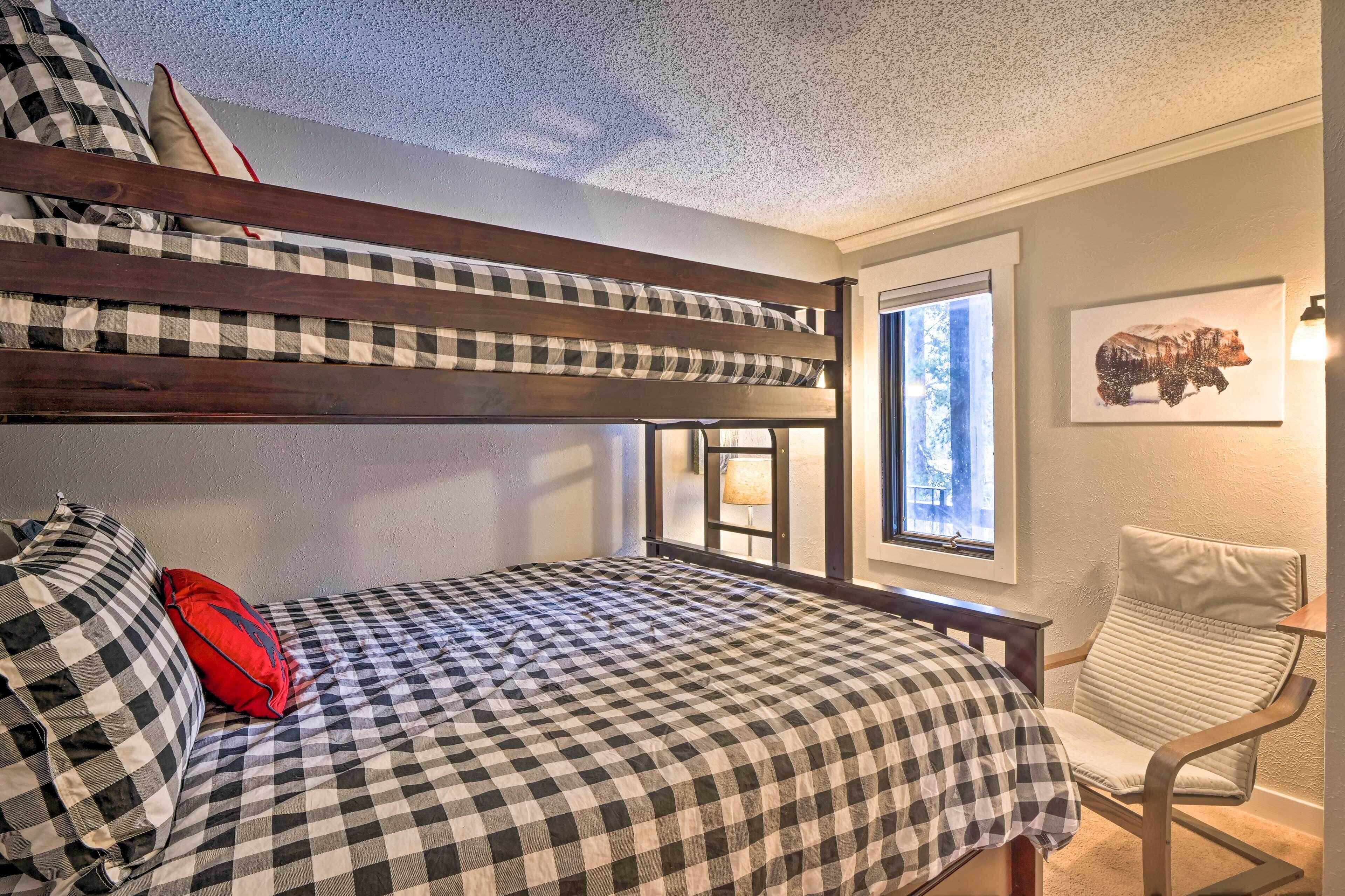 A bunk bed with checkered bedding, a red pillow, a window with a view of trees, and a chair with a light fixture.
