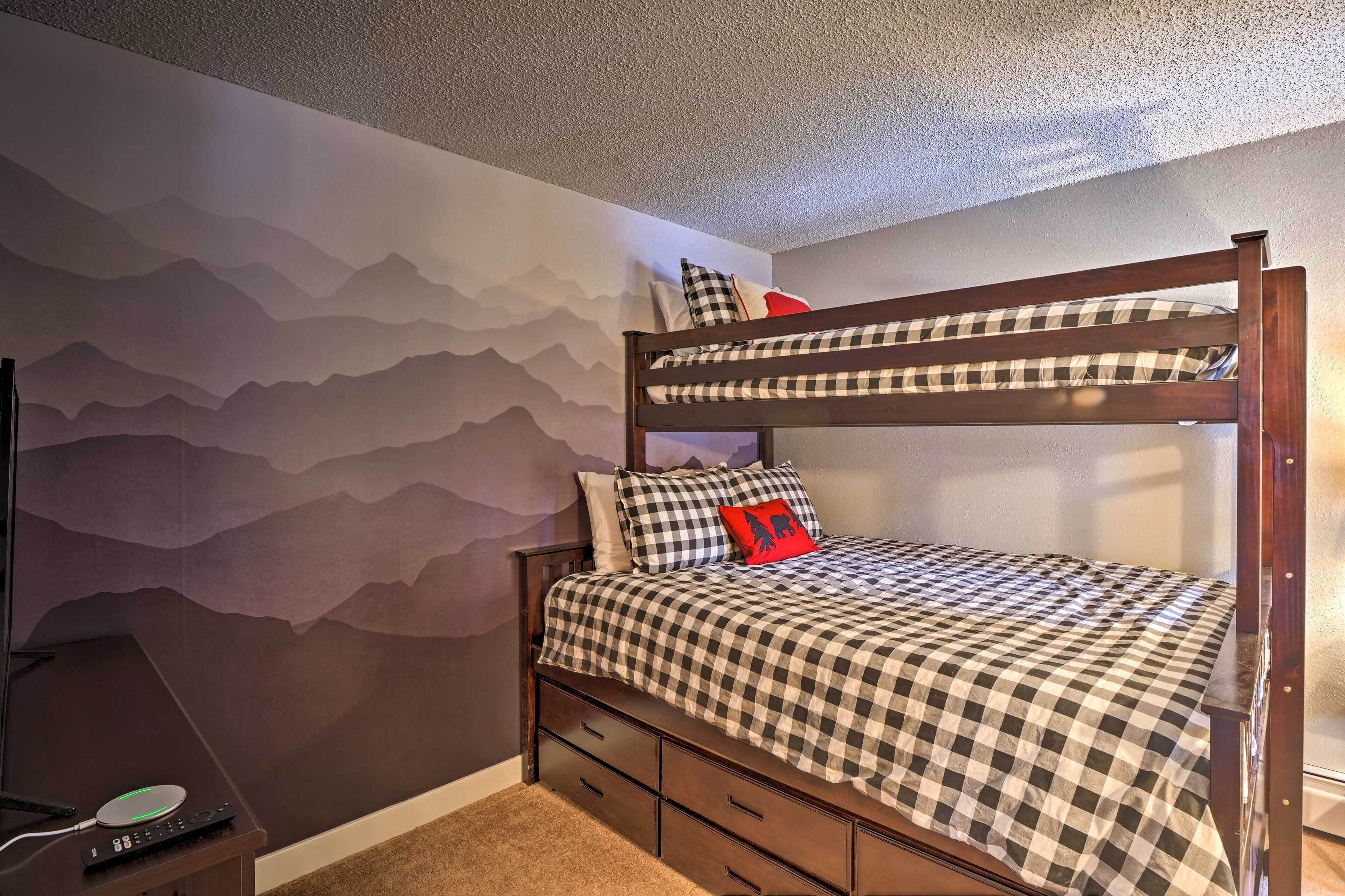 A bunk bed with a checkered bedspread and pillows, a small desk with a lamp, and a wall with a mountain design.