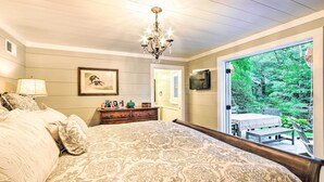 A bedroom with a large bed, a chandelier, a dresser, a television, and a sliding glass door leading to a balcony with a view of greenery.