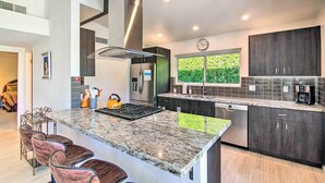 A modern kitchen with dark cabinetry, a large island with a built-in stove, stainless steel appliances, and a window with a view of greenery. - Palm Springs Oasis w/ Pool & Golf Course Views! (Palm Springs)