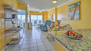 A bright living room with yellow walls, a dining area with a fruit bowl, and a balcony with outdoor furniture.