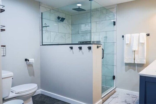 Moderni kylpyhuone, jossa on lasiseinällä varustettu suihku, wc ja pesuallas, jonka päällä on pyyhkeitä.