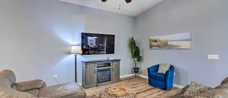 Een woonkamer met een plafondventilator, een open haard, een blauwe fauteuil en een grote plant.