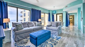 A spacious living room with two sofas, a blue ottoman, and a large window with curtains. - Chic Panama City Beach Condo w/ Pool & Gulf Views (Panama City Beach)