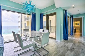 A modern dining area with a glass table and chairs, a view of the ocean, and a balcony with blue curtains. - Chic Panama City Beach Condo w/ Pool & Gulf Views (Panama City Beach)