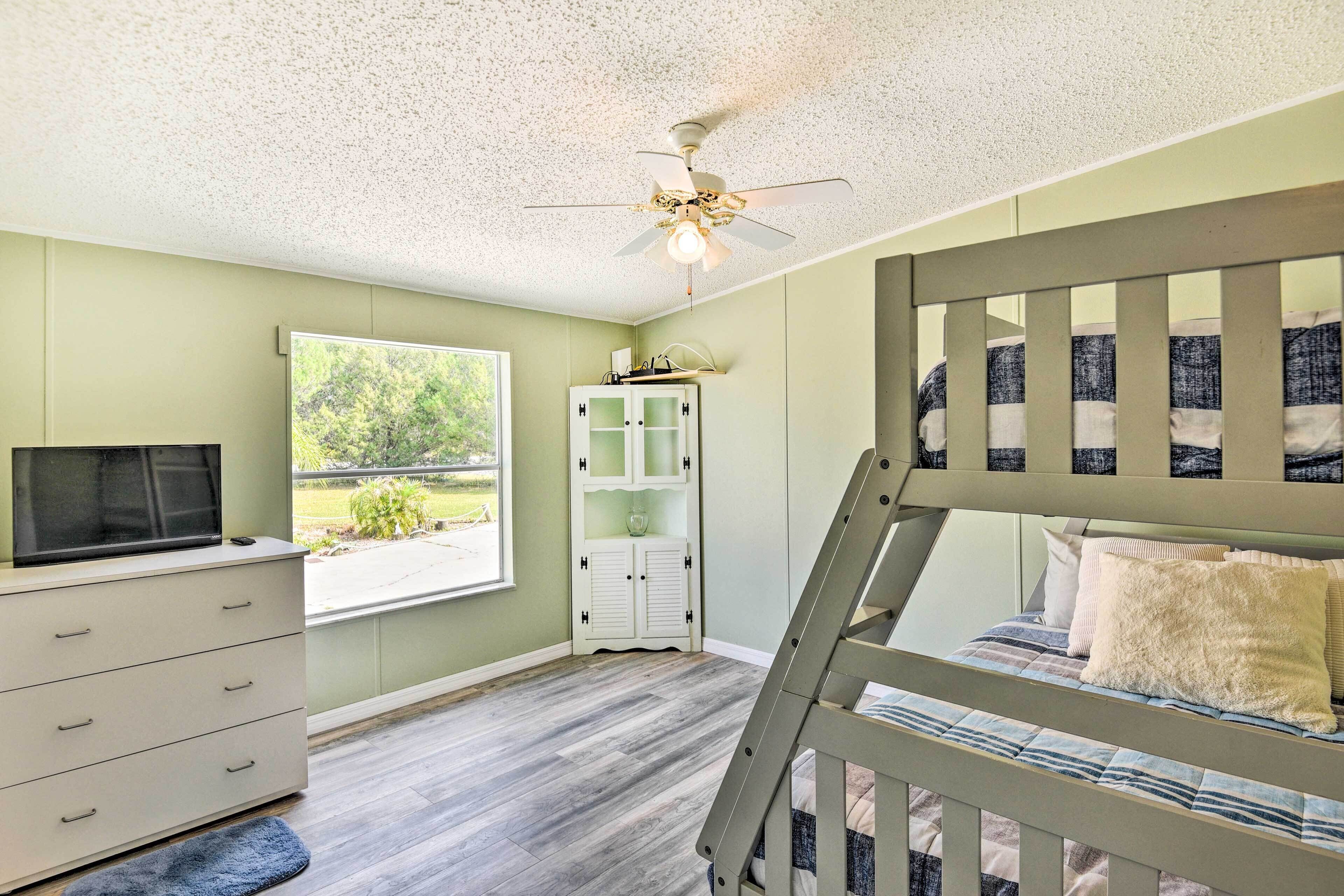 A bedroom with a bunk bed, a white dresser, a television, a ceiling fan, and a window with a view of greenery.