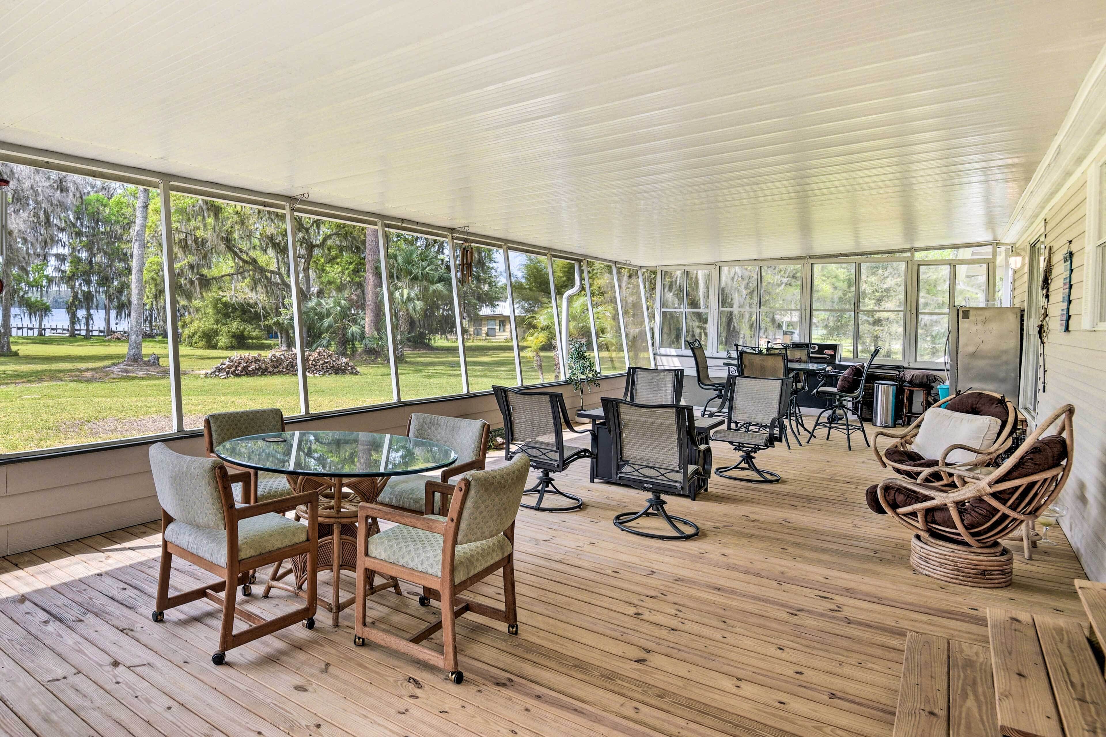 A covered outdoor area with a glass table and chairs, surrounded by a wooden deck and large windows offering views of a grassy area and trees.