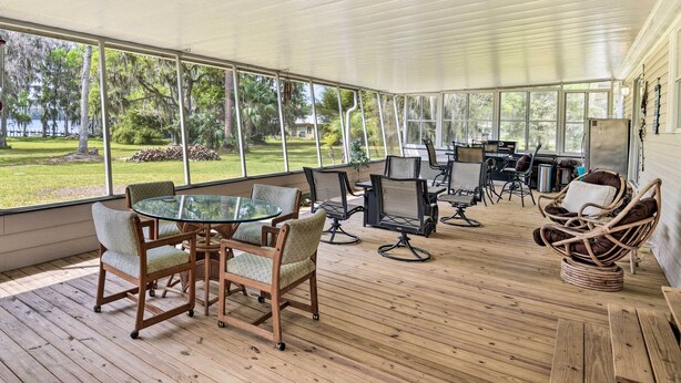 A covered outdoor area with a glass table and chairs, surrounded by a wooden deck and large windows offering views of a grassy area and trees. - Waterfront Home w/ Boat Dock & Large Porch! (Georgetown)