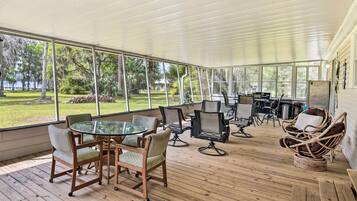 A covered outdoor area with a glass table and chairs, surrounded by a wooden deck and large windows offering views of a grassy area and trees.