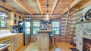 A rustic cabin kitchen with wooden cabinets, a central island, a stone fireplace, and a view through the windows. - Restored Buchanan Log Cabin on 9-mile Creek! (Buchanan)
