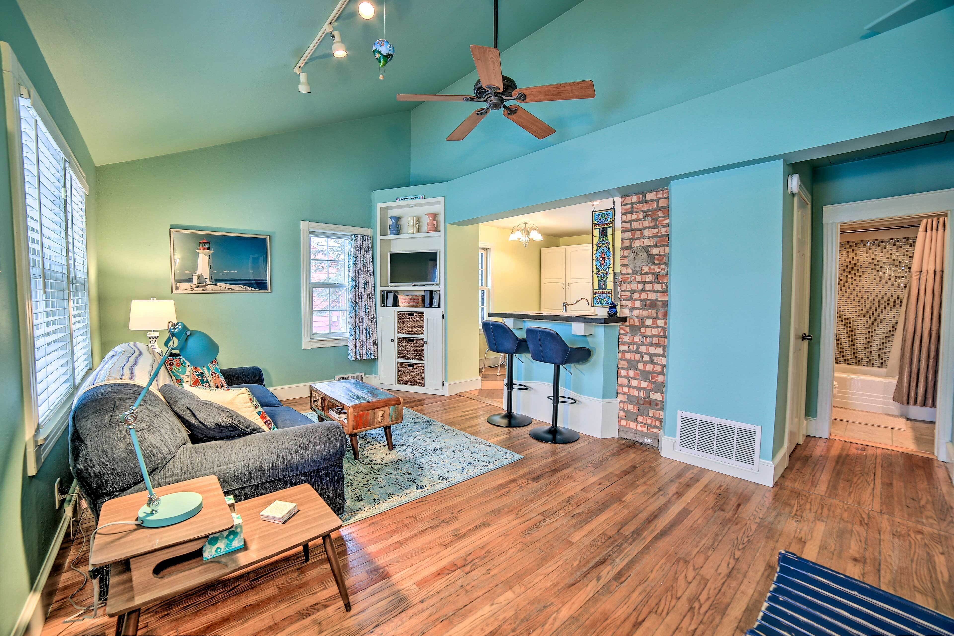 A living room with a sofa, coffee table, and a ceiling fan. There is a kitchen area with a brick wall and a window.