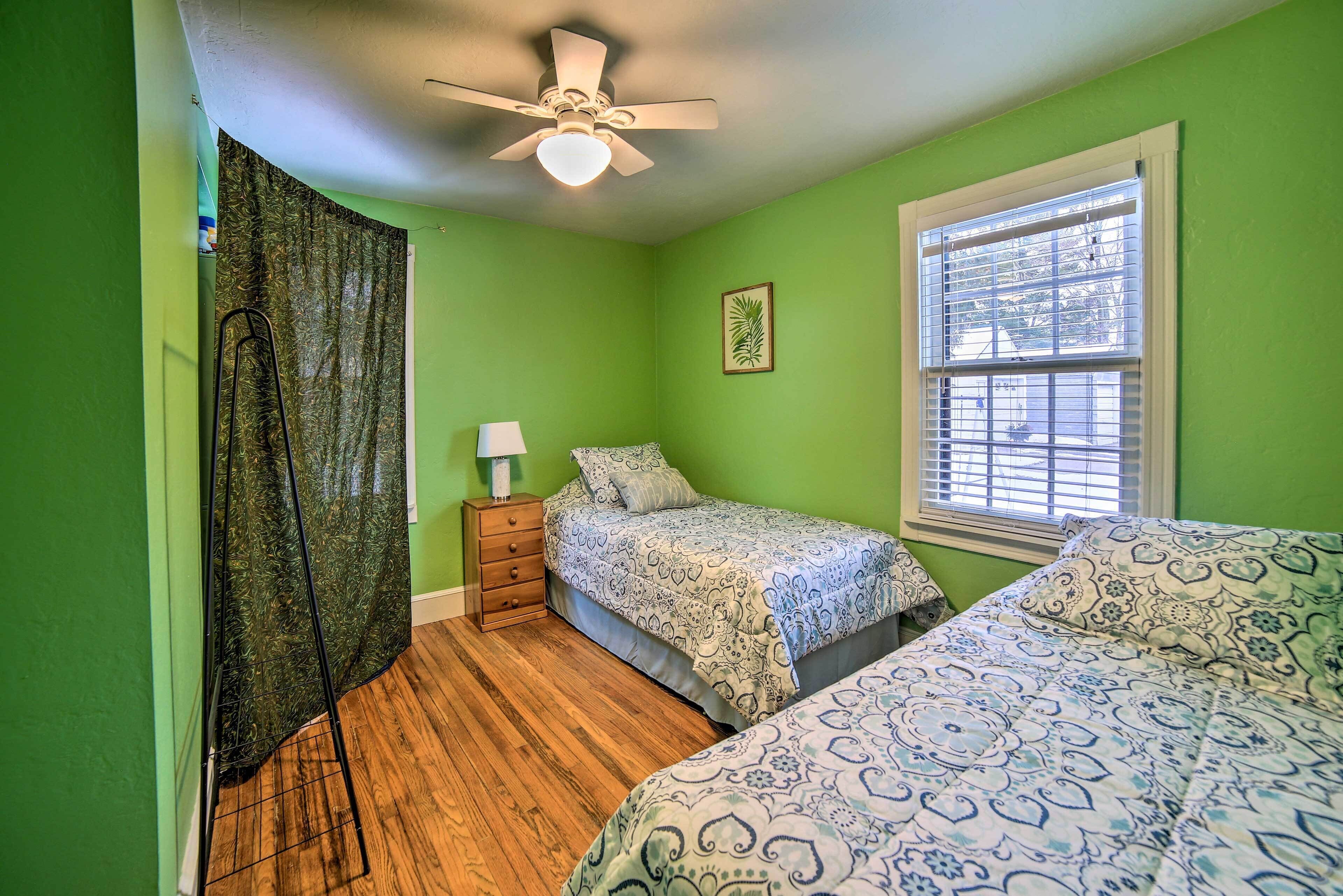 A bedroom with two beds, a ceiling fan, a window with blinds, a nightstand with a lamp, and a framed picture on the wall.