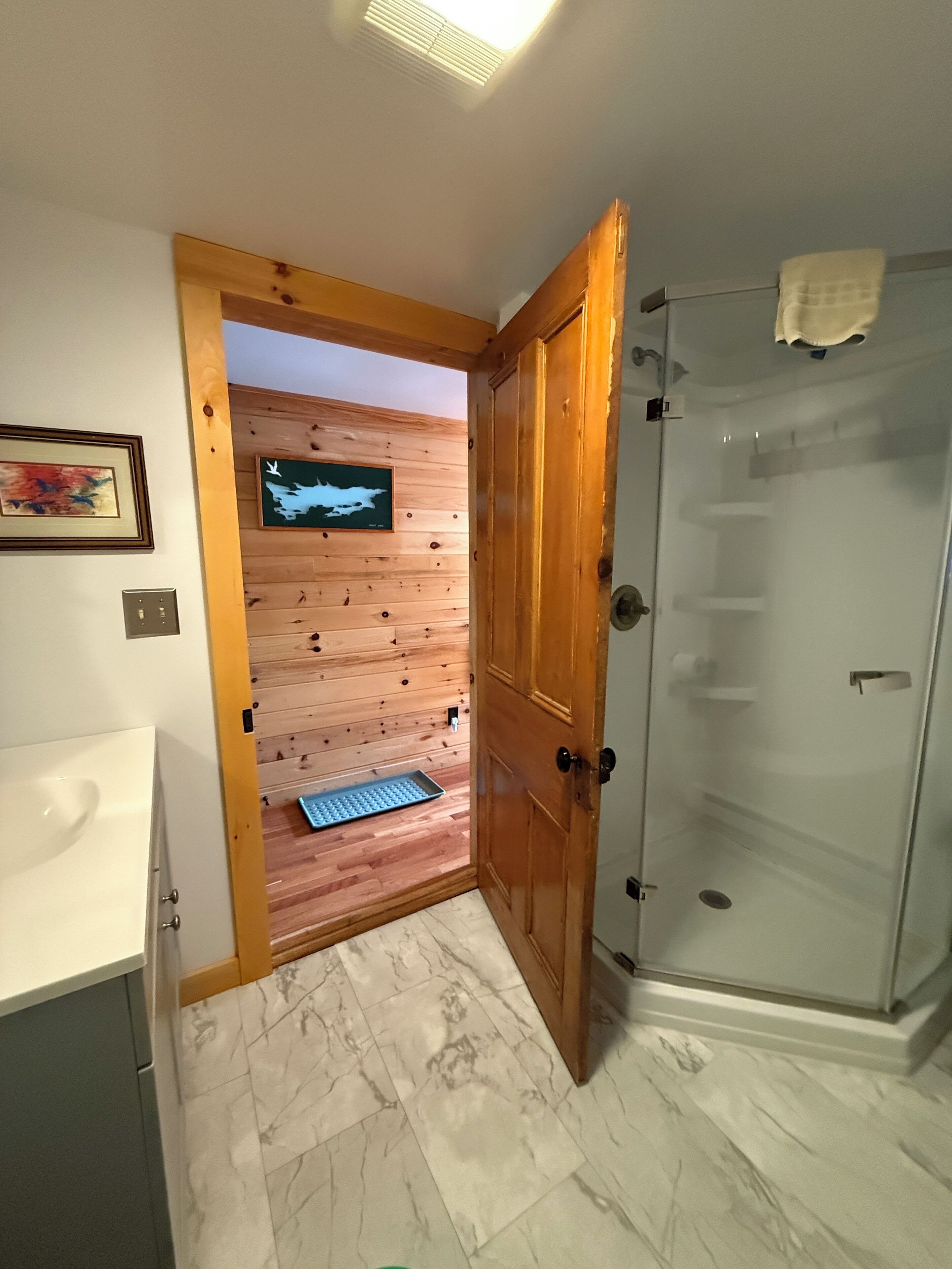 A bathroom with a shower, a sink, and a wooden door leading to a room with a wooden floor and a wall-mounted TV.