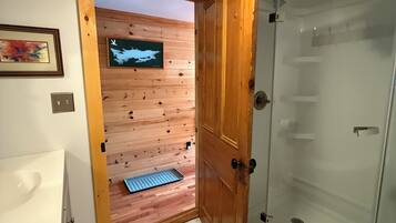 A bathroom with a shower, a sink, and a wooden door leading to a room with a wooden floor and a wall-mounted TV.