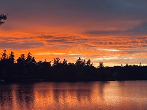 Un coucher de soleil sur un lac paisible, avec les silhouettes d’arbres et une rive lointaine.