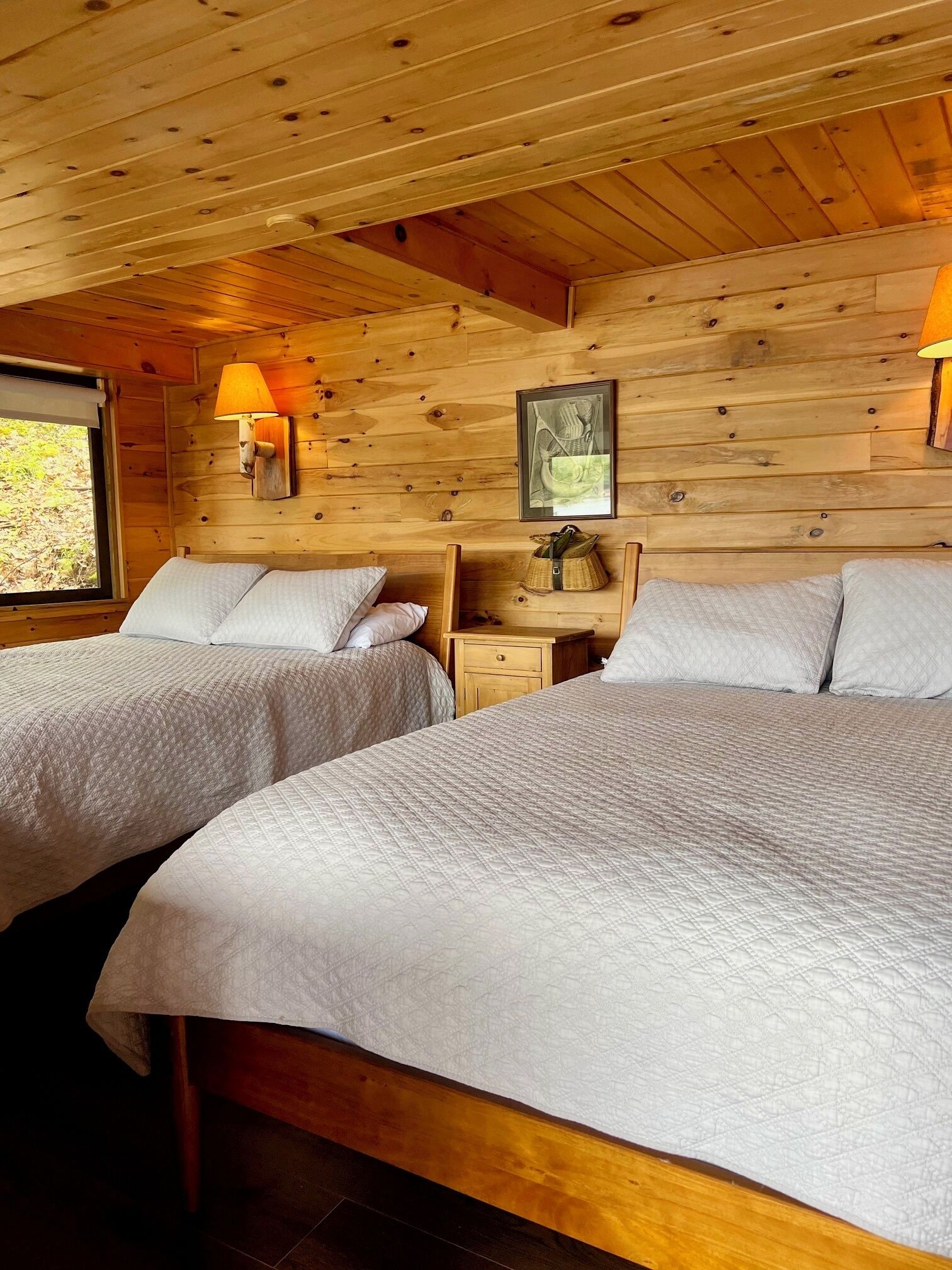 A wooden cabin room with two beds, a nightstand, a lamp, and a framed picture on the wall.