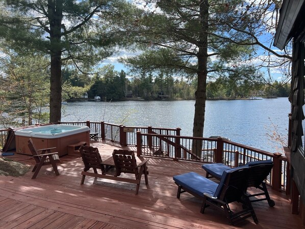 Une terrasse en bois avec un jacuzzi, des chaises longues et une vue sur un lac entouré d’arbres.