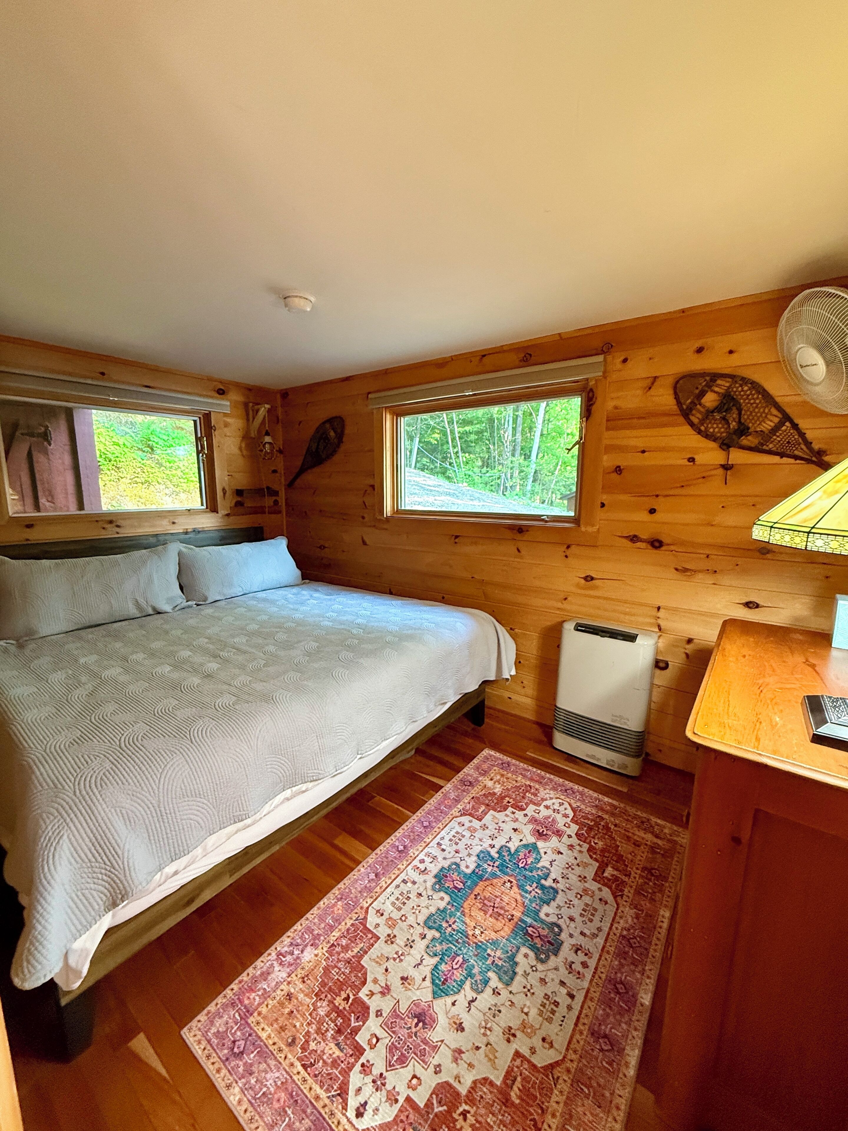 Ein Schlafzimmer einer Holzhütte mit einem Bett, zwei Fenstern, einem Heizgerät, einem Ventilator und einem Teppich.