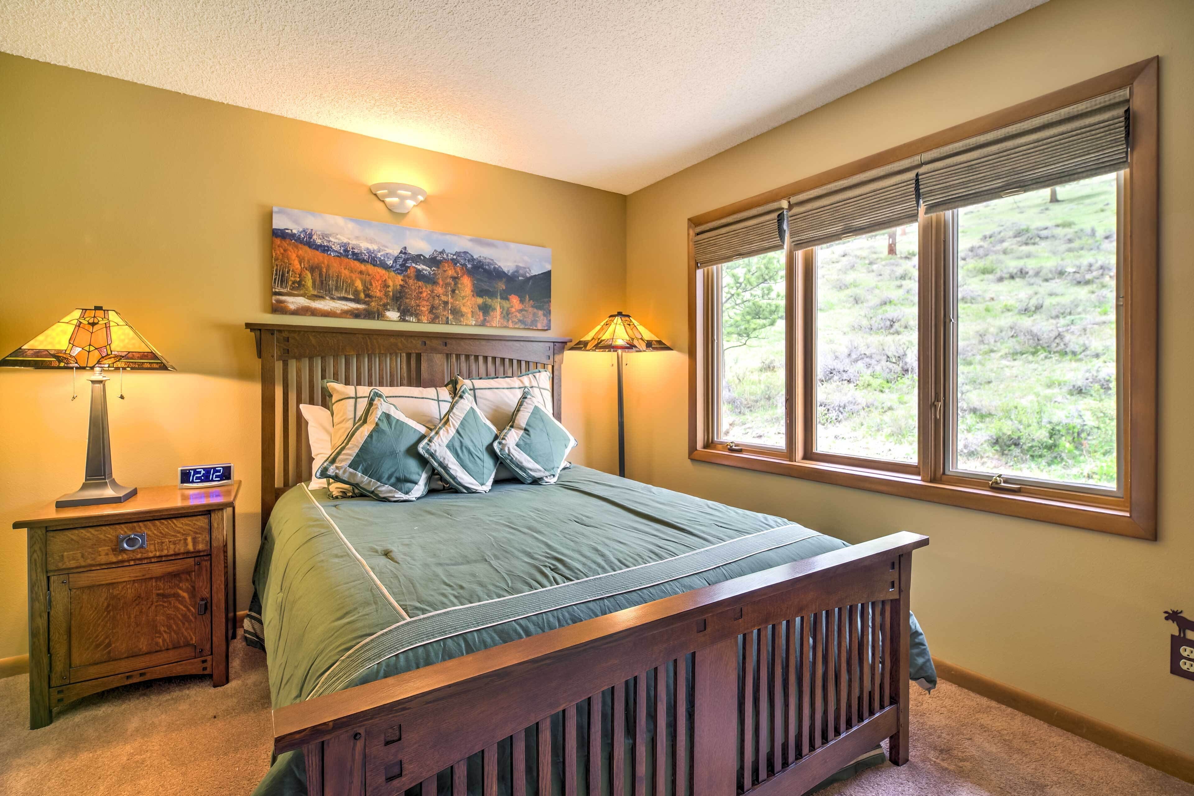 A bedroom with a bed, two bedside lamps, a nightstand, a painting, and a window with a view of a landscape.