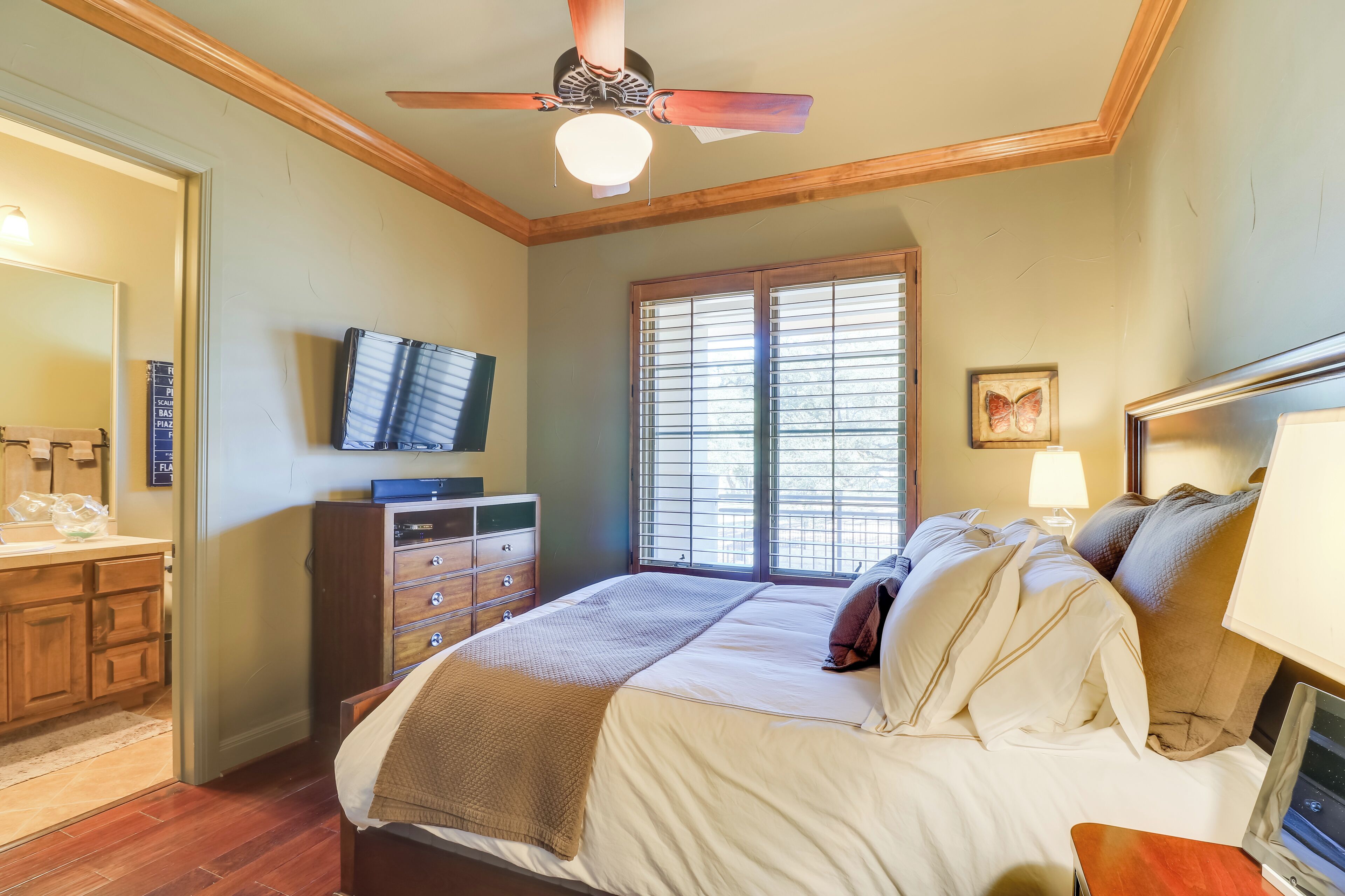 Un dormitorio con cama, ventilador de techo, televisor, ventana con persianas, cómoda y mesita de noche.