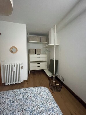 A bedroom with a bed, a radiator, a white dresser, a shelf with boxes, and a hanging rack.