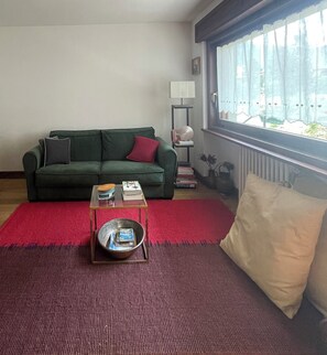 Ett vardagsrum med en grön soffa, en röd matta, ett soffbord i glas och ett fönster med gardiner.
