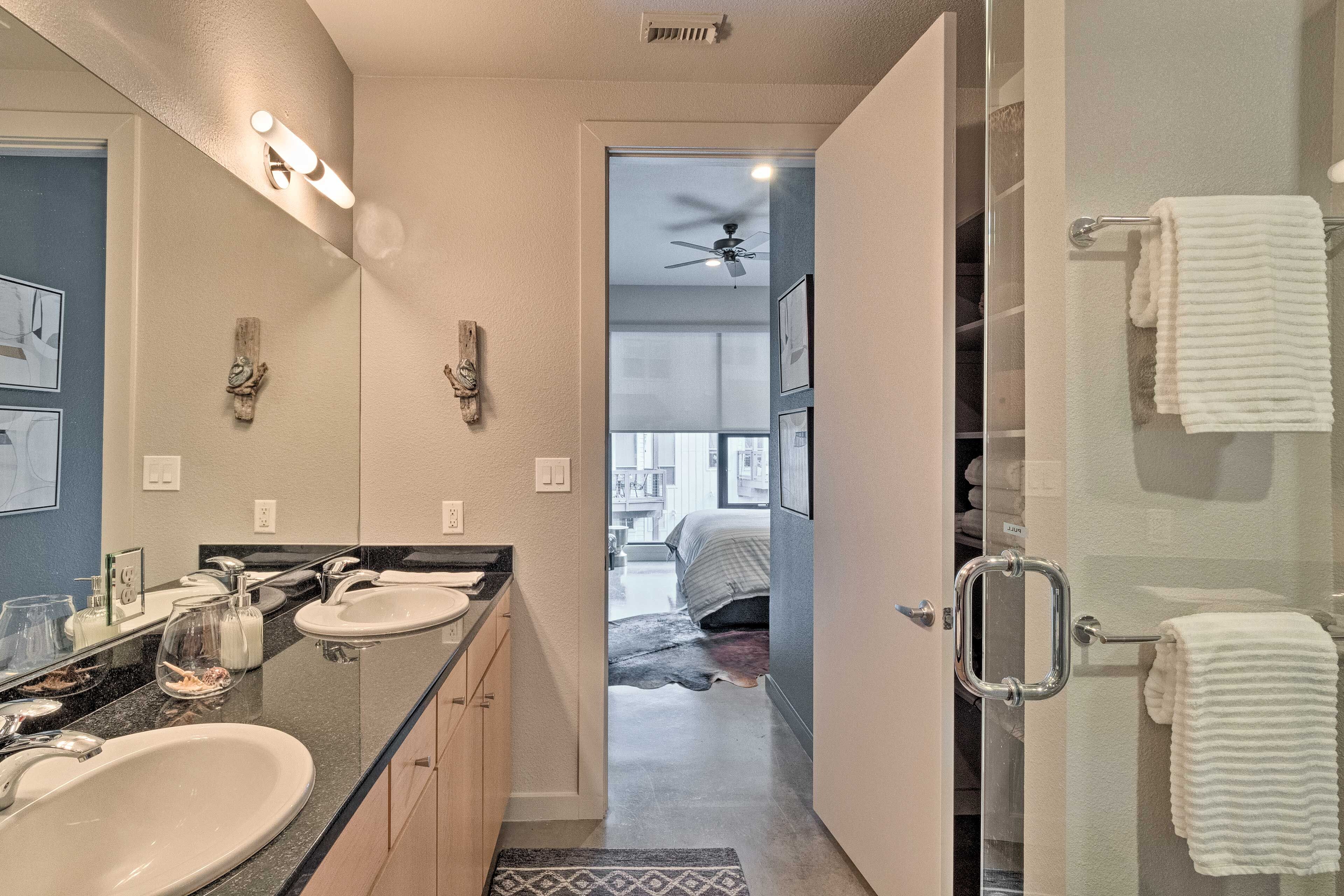A modern bathroom with two sinks, a glass shower enclosure, and a visible bedroom in the background.