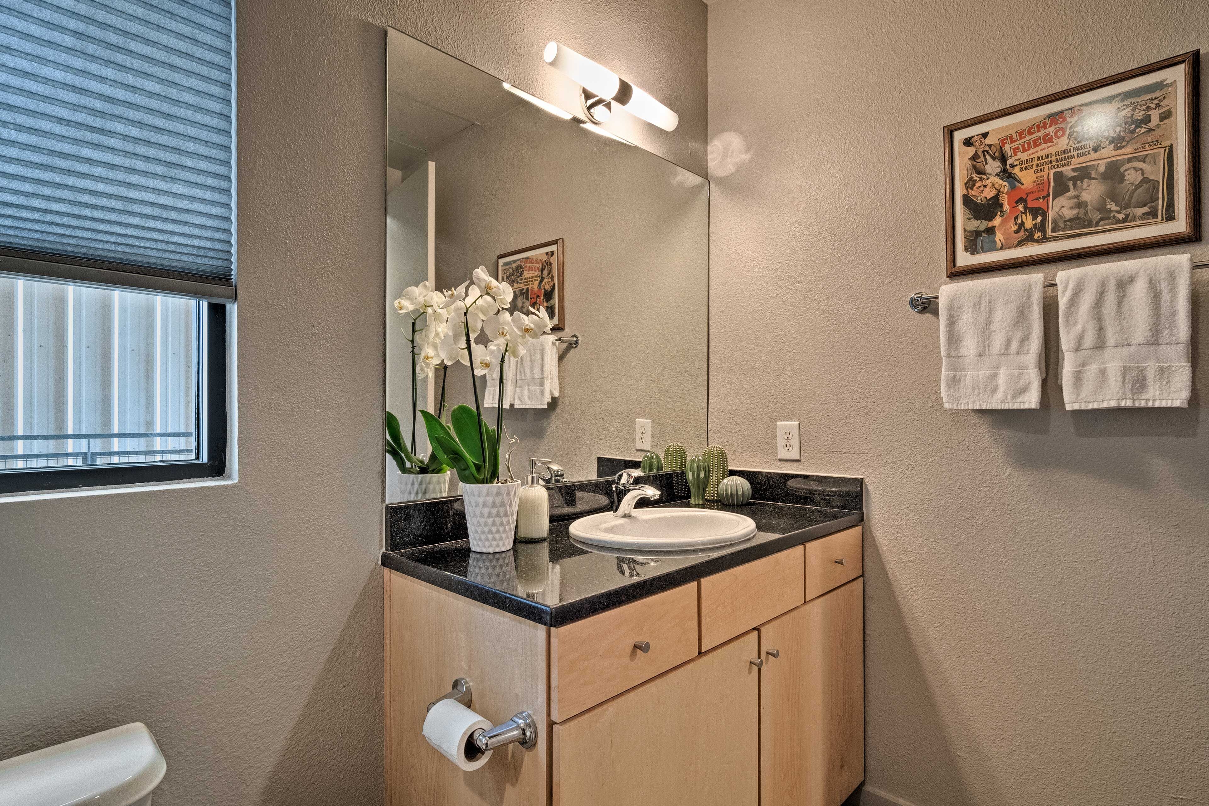 Een badkamer met een wastafel, spiegel en toilet. Er is een raam met jaloezieën, een handdoekhouder en een lijst met een foto aan de muur.