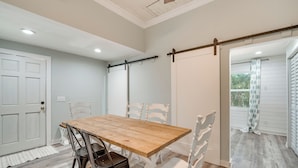 Un comedor con una mesa de madera y sillas blancas, una puerta blanca y vistas al exterior a través de una ventana.