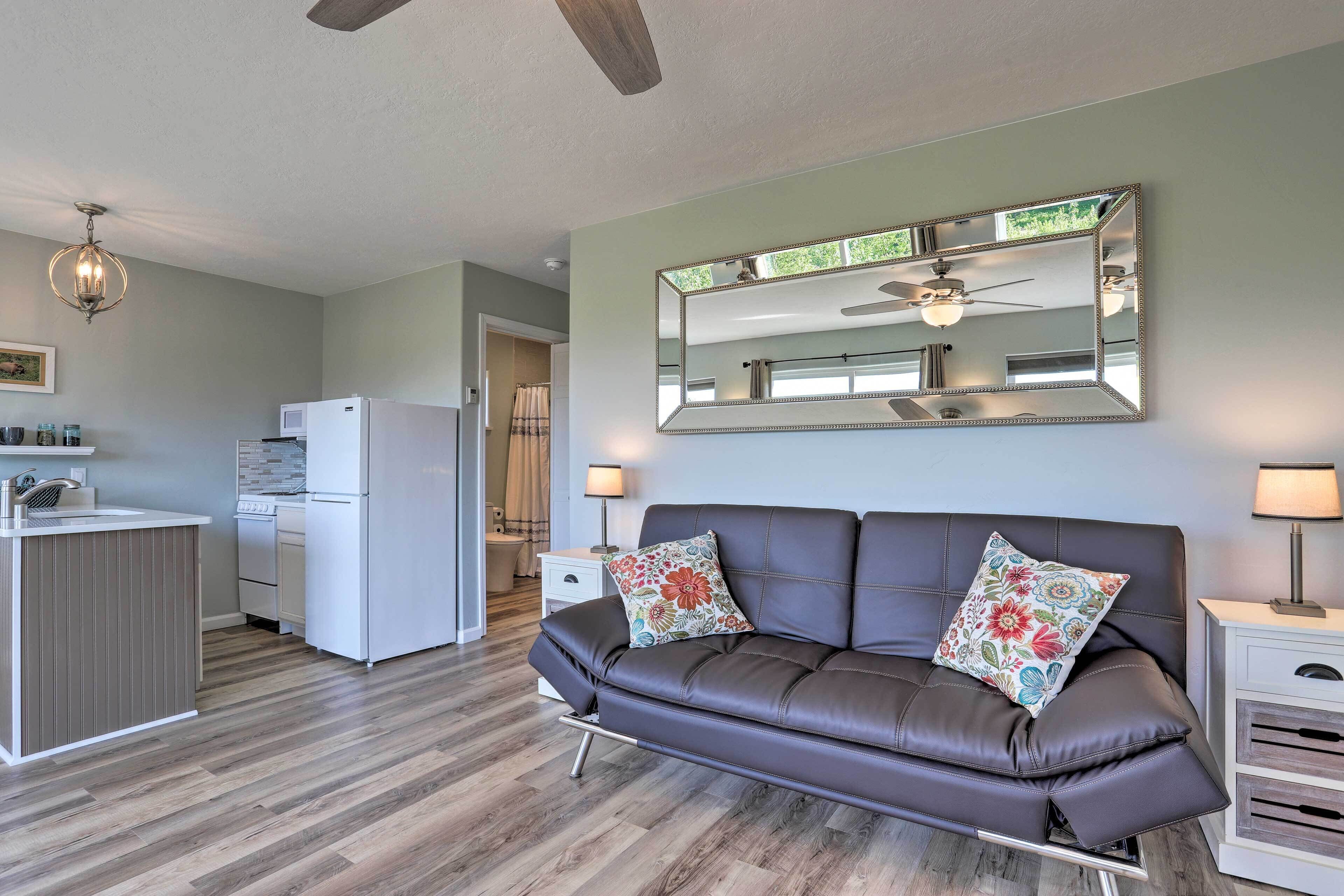 A modern living room with a dark leather sofa, a large mirror, and a kitchen area with white appliances.