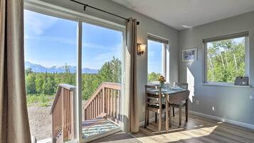 A room with large sliding glass doors leading to a balcony with a wooden railing, a dining table with chairs, and a view of mountains and trees.