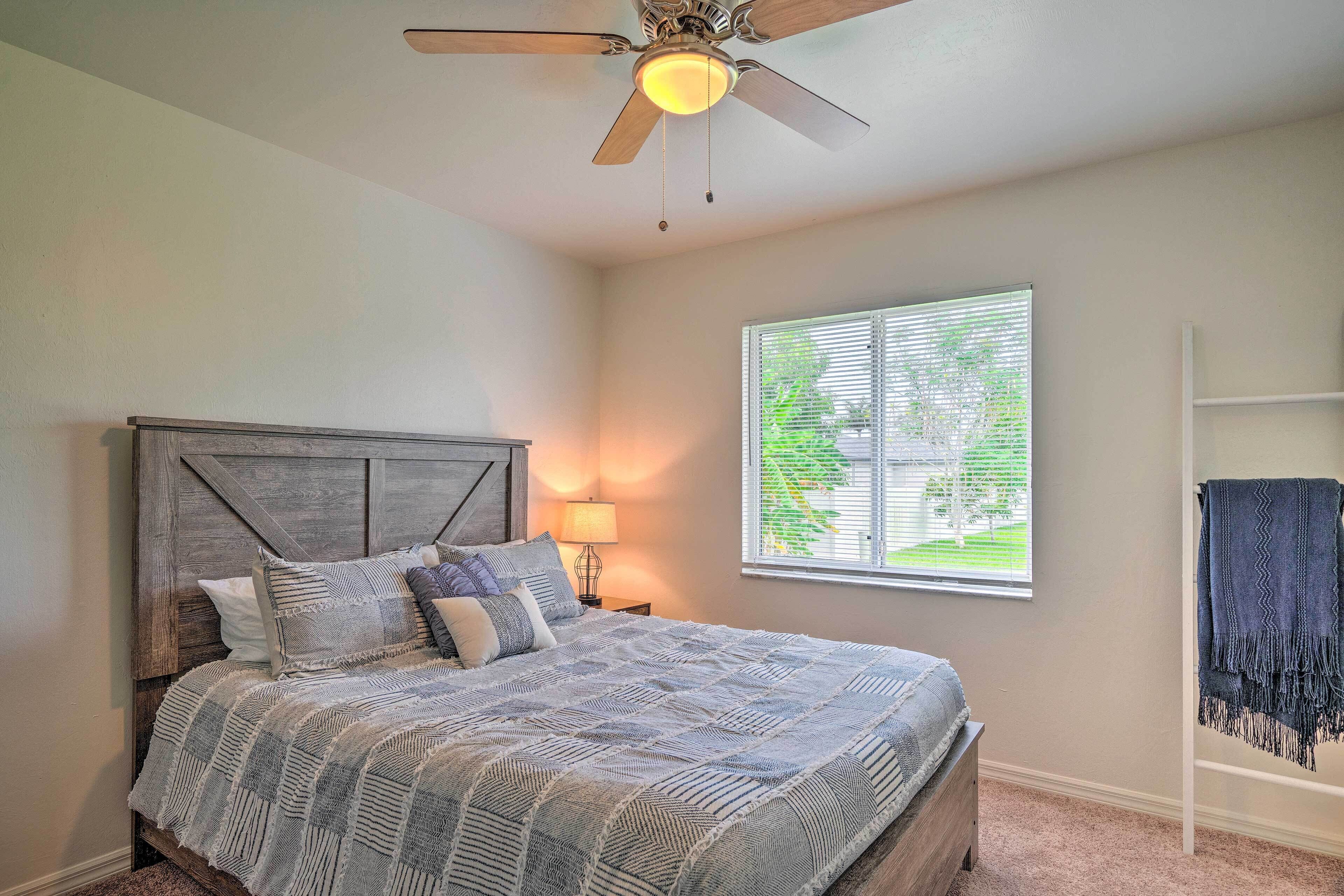 A bedroom with a bed, a ceiling fan, a window with blinds, and a nightstand with a lamp.