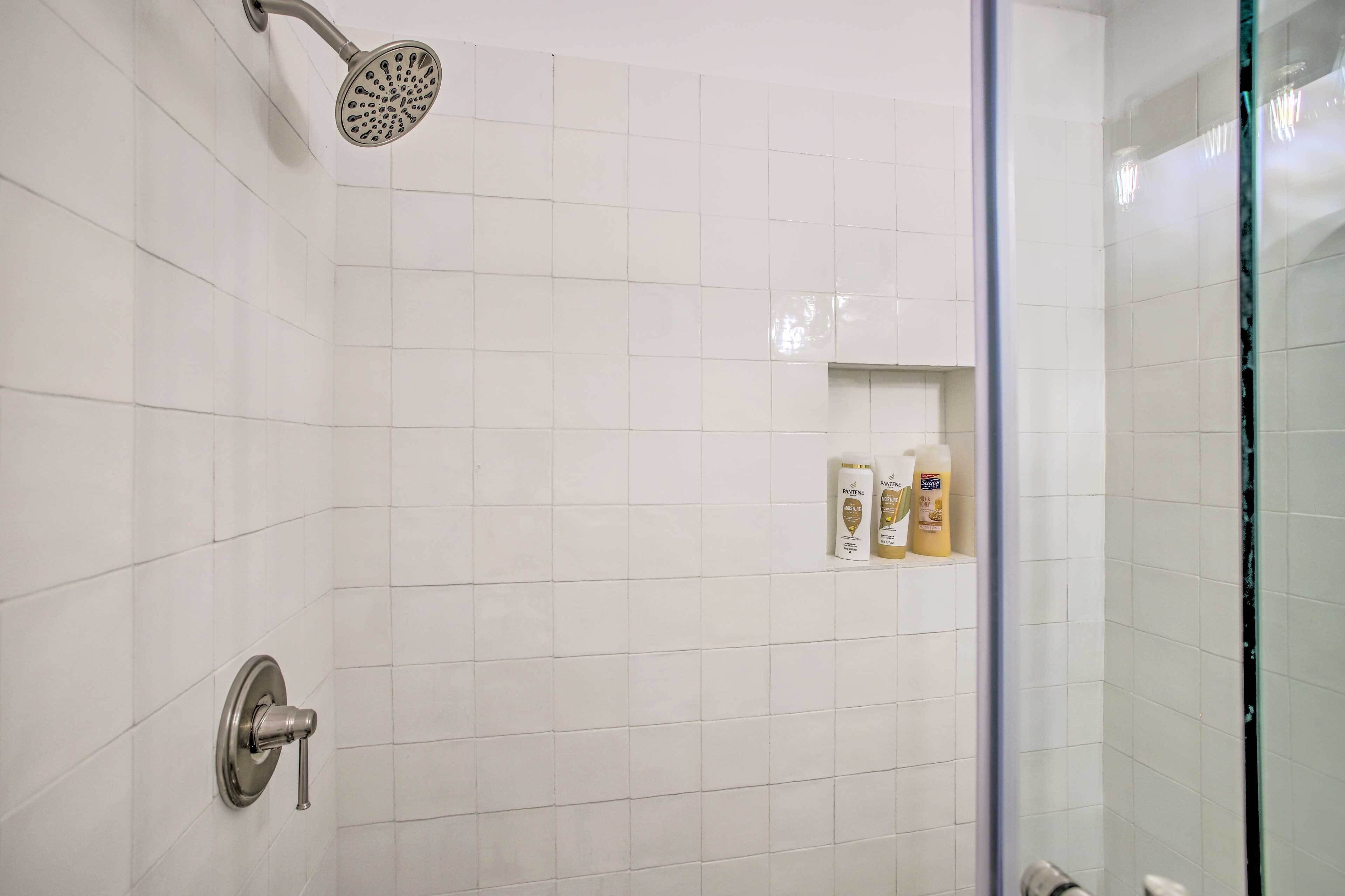 A bathroom with white tiles, a showerhead, a toilet, and a shelf with toiletries.