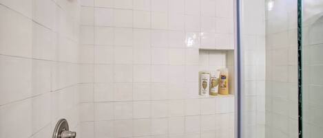A bathroom with white tiles, a showerhead, a toilet, and a shelf with toiletries.
