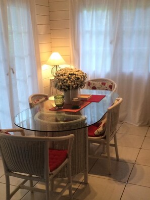 Un comedor con una mesa de vidrio rodeada de sillas de mimbre con cojines rojos. Un jarrón con flores y una lámpara están sobre la mesa.