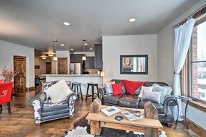 A spacious living room with a leather sofa, a wooden coffee table, and a red chair. A kitchen is visible in the background with modern appliances and a bar area. - Main St Bozeman Condo - Walk to Parks & Eateries! (Bozeman)