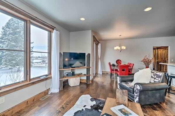 A spacious living room with a large window, a flat-screen TV, a wooden floor, a leather sofa, and a dining area with red chairs. - Main St Bozeman Condo - Walk to Parks & Eateries! (Bozeman)