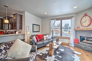 A living room with a fireplace, a large clock, and a view of a snowy landscape through the window. - Main St Bozeman Condo - Walk to Parks & Eateries! (Bozeman)
