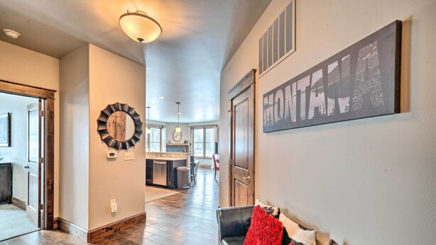 A spacious living area with a dark leather sofa, a red throw pillow, and a round mirror on the wall. A kitchen is visible in the background, and there is a large framed picture on the wall. - Main St Bozeman Condo - Walk to Parks & Eateries! (Bozeman)