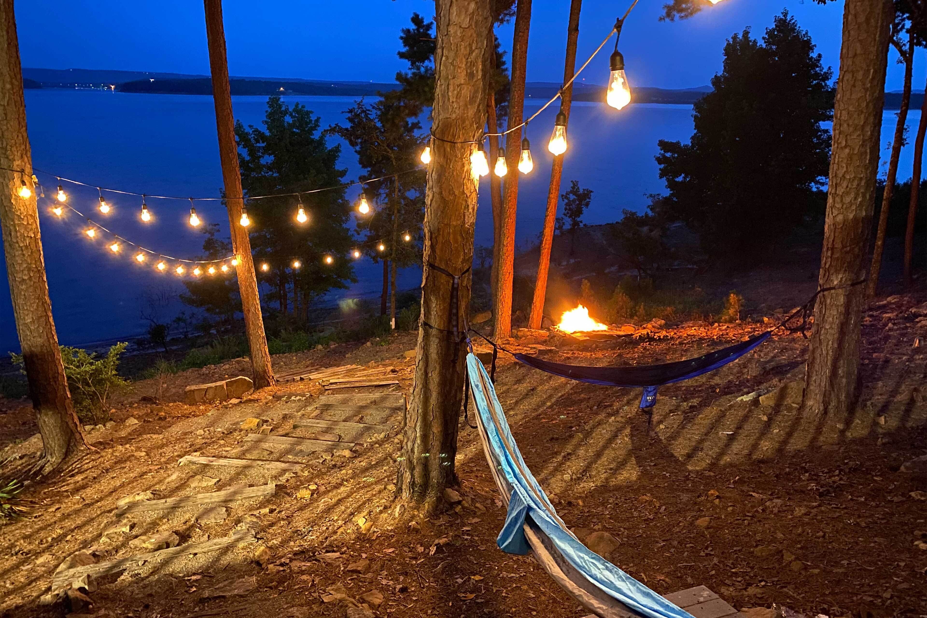 A hammock tied between trees, a campfire, and string lights over a forested area near a body of water.