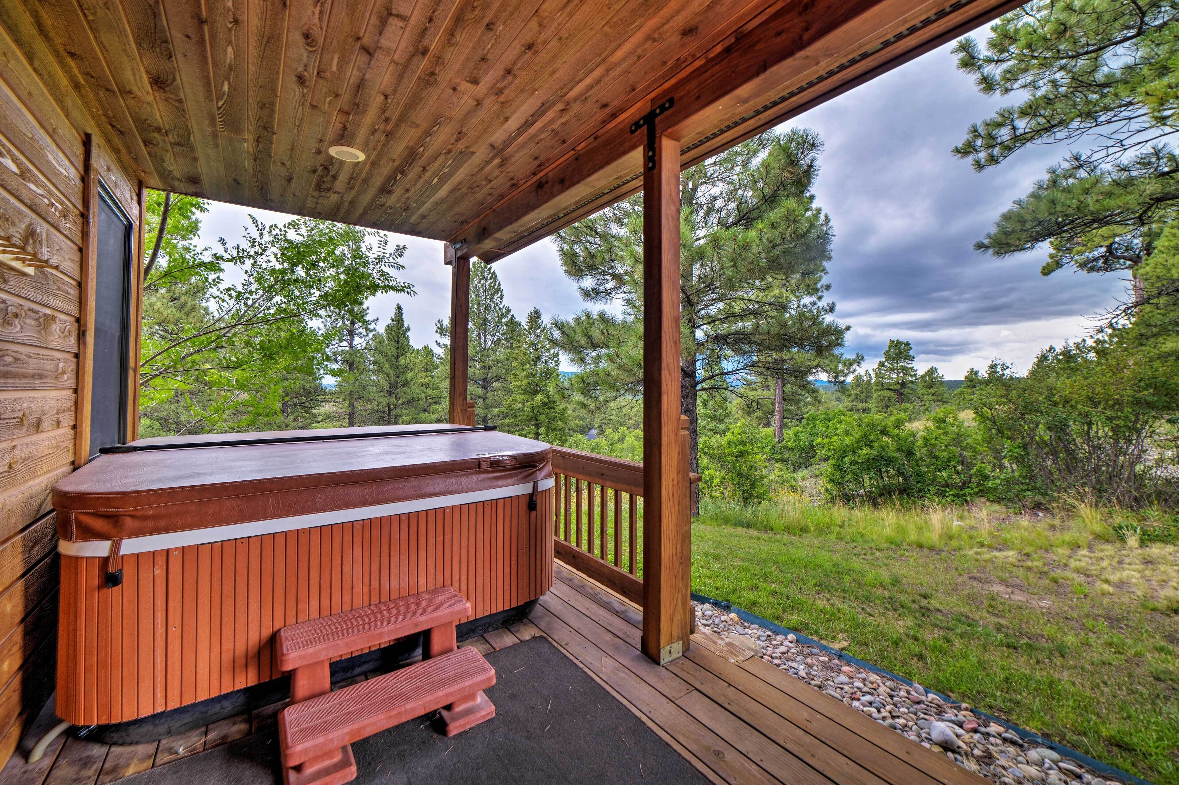 A wooden deck with a hot tub and benches, surrounded by trees and a grassy area.